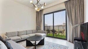 Un salon moderne avec un canapé d’angle, une table basse et un téléviseur. La pièce est dotée de grandes fenêtres avec des rideaux et offre une vue sur les bâtiments environnants.