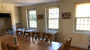 Uma área de refeições com mesa e cadeiras de madeira, uma cozinha com fogão e forno, e duas janelas com detalhes artísticos.