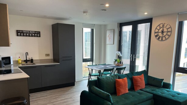 Ein modernes Wohnzimmer mit einem grünen Ecksofa, einem Esstisch mit Stühlen, einer Küchenzeile und einem großen Fenster mit Jalousien.