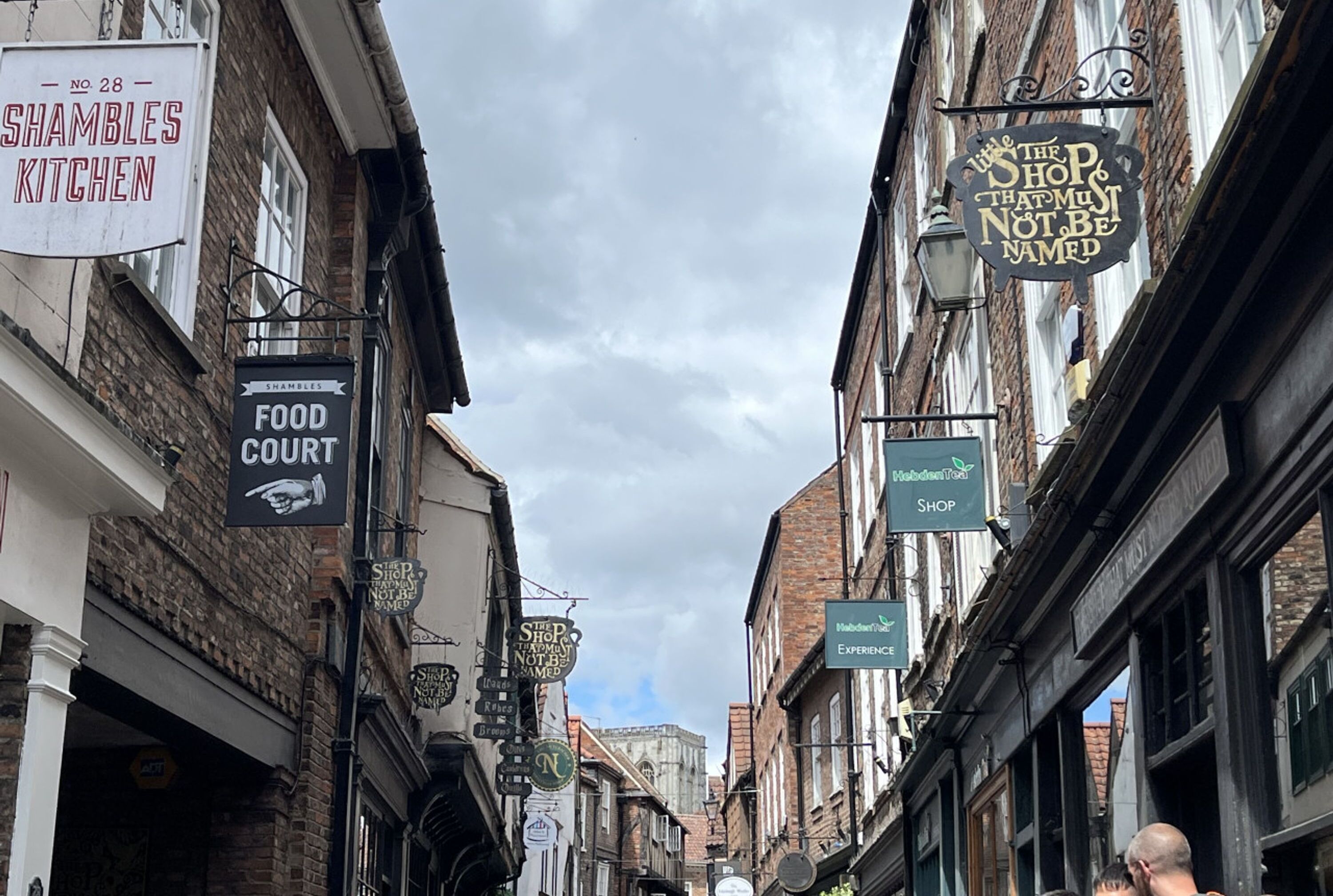 Una calle estrecha con diversas tiendas y letreros, incluyendo una zona de comida y un restaurante llamado 'Shambles Kitchen'.