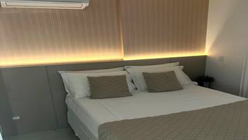 Una cama bien hecha con cubrecolcha acolchado, una mesita de noche con lámpara y un aire acondicionado de pared.