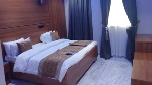 A modern bedroom with a wooden headboard, a large bed with white and brown bedding, a wooden nightstand, and a window with dark curtains.