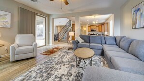 Un salon spacieux avec un canapé d’angle gris, un fauteuil blanc et une table basse. Un ventilateur de plafond et un escalier sont visibles à l’arrière-plan.