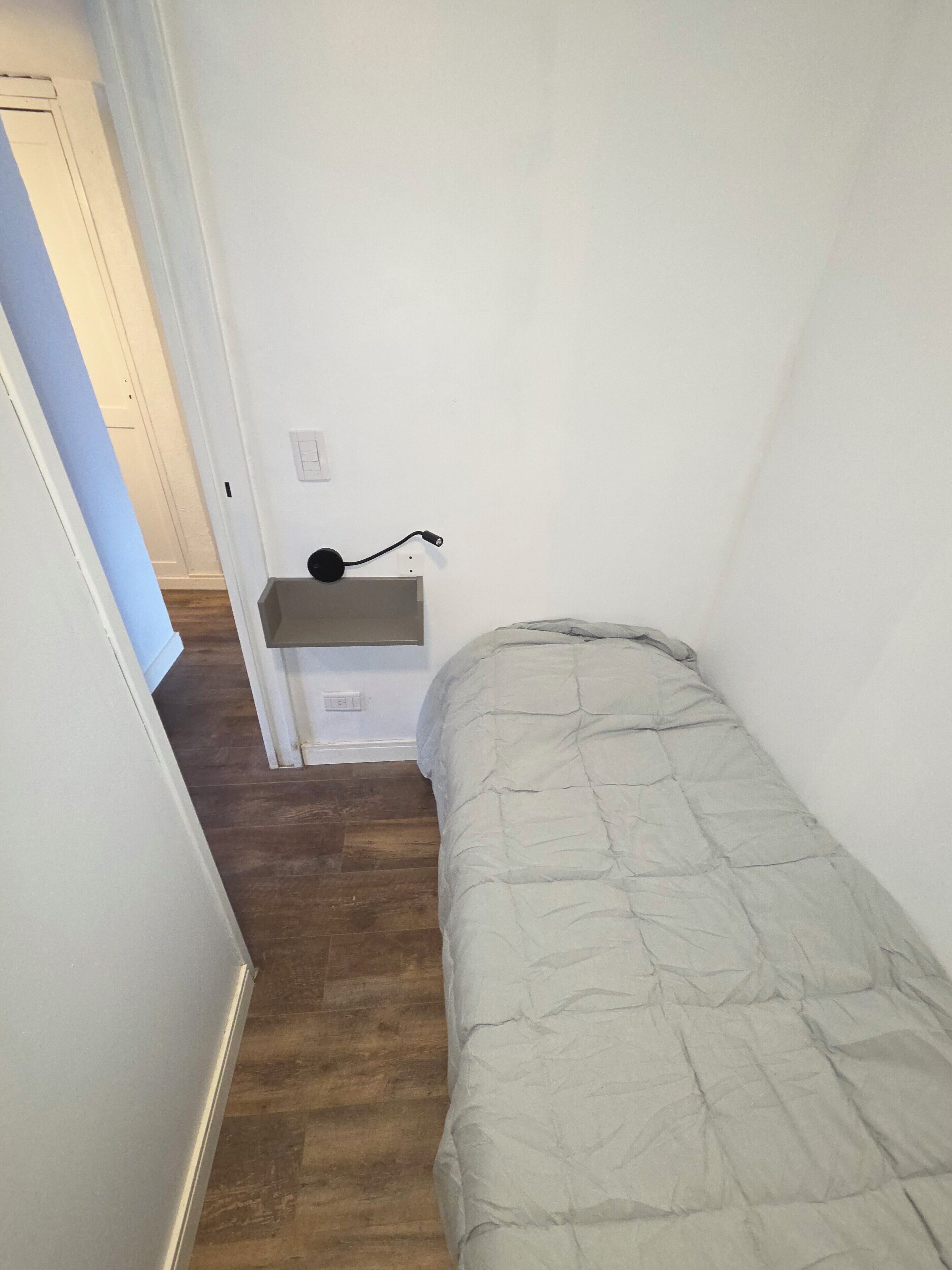 A narrow hallway with a bed on the right, a small desk with a lamp, and a door at the end.