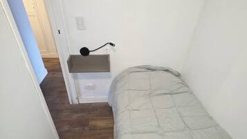 A narrow hallway with a bed on the right, a small desk with a lamp, and a door at the end.