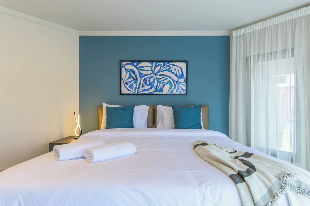 Una cama bien hecha con sábanas blancas, un cuadro abstracto azul en la pared y una ventana con cortinas de gasa.