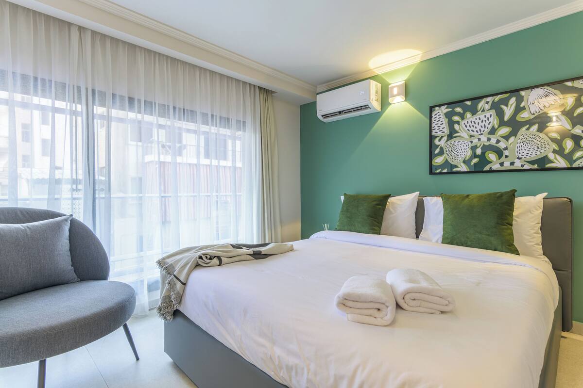 Habitación de hotel con cama, un sillón gris, un ventanal con cortinas traslúcidas, aire acondicionado instalado en la pared y una obra de arte floral.