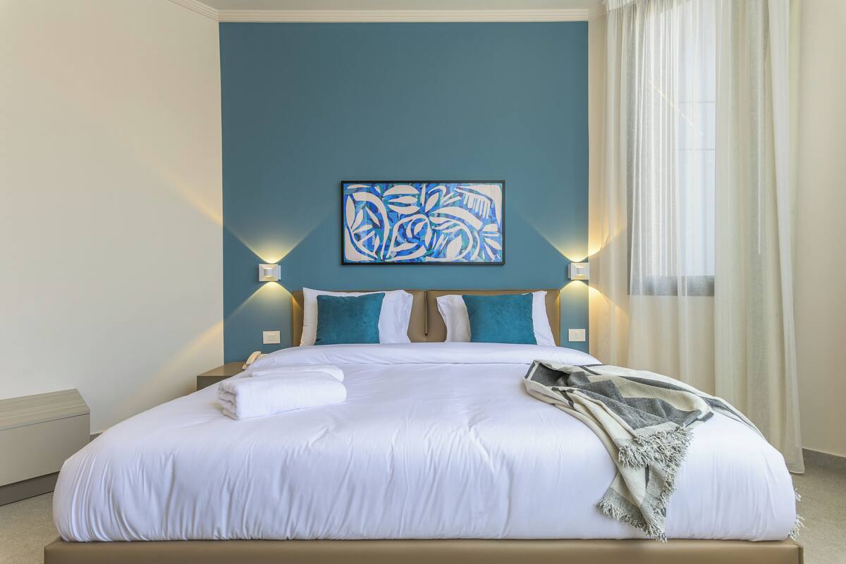 Una cama bien hecha con sábanas blancas, dos almohadas y una colcha. En la pared sobre la cama, un cuadro abstracto azul.