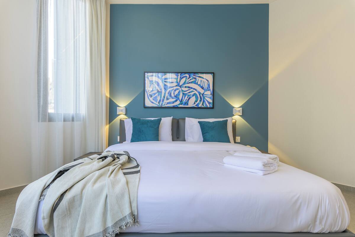 Una cama bien hecha con sábanas blancas y almohadas turquesa, un cuadro abstracto grande en la pared y dos lámparas fijadas a la pared.