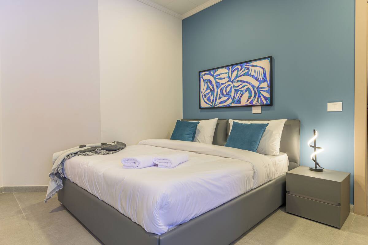 Una cama bien tendida con sábanas blancas y dos almohadas, una mesita de noche con una lámpara y un cuadro abstracto azul en la pared.