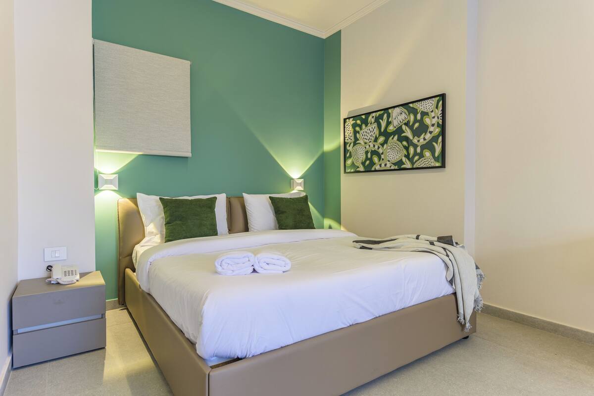 Una cama bien hecha con ropa blanca, una lámpara de noche y una mesita de noche con un teléfono.