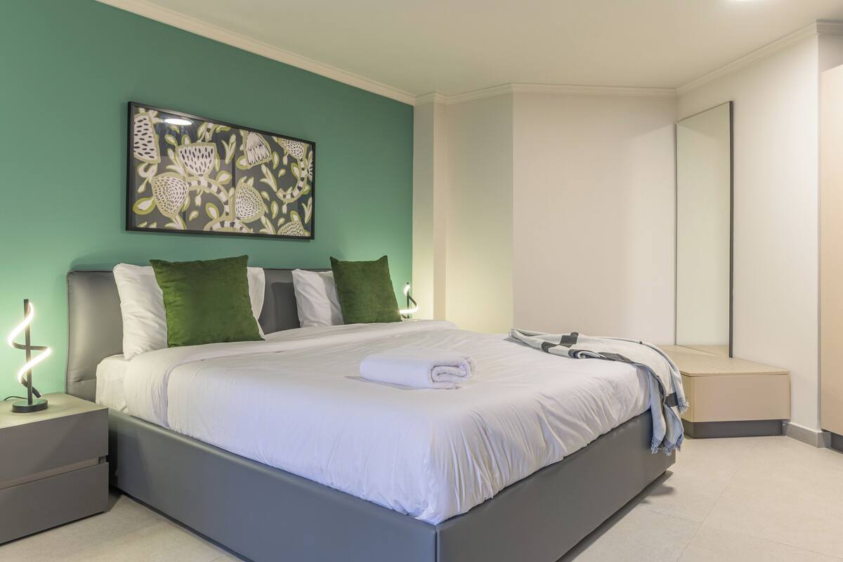 Una cama bien hecha con sábanas blancas y almohadas verdes, una mesita de noche con lámpara, y un espejo grande en la pared.
