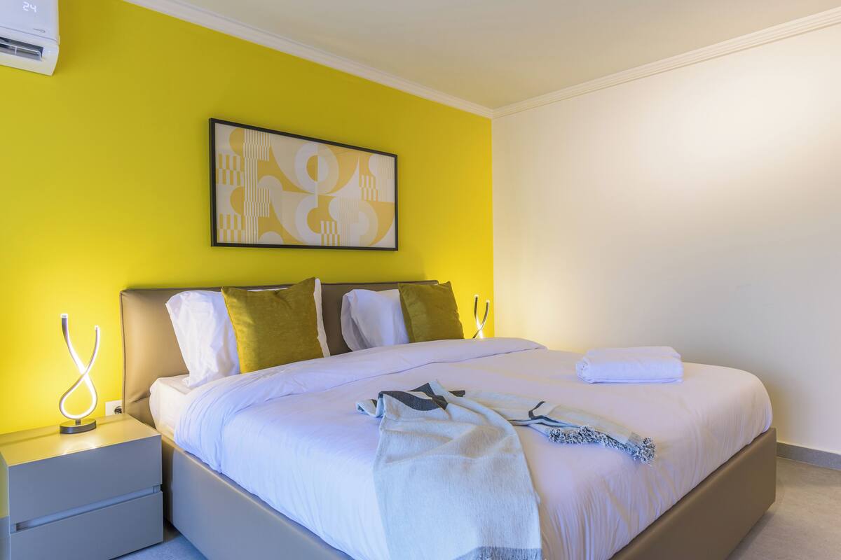 Habitación de hotel con cama, mesitas de noche, aire acondicionado de pared y una obra de arte abstracta enmarcada.