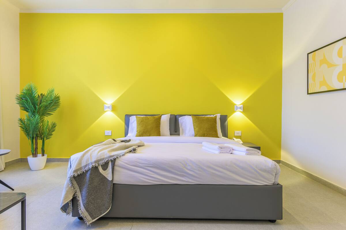 Un dormitorio moderno con una pared de acento amarilla, una cama con ropa de cama blanca, dos lámparas de noche y una planta en maceta.