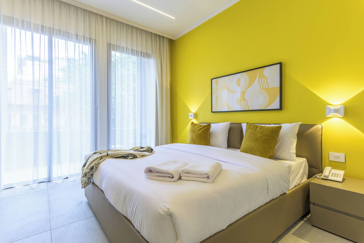 Habitación de hotel con una cama grande, una mesita de noche con teléfono y una ventana con cortinas traslúcidas.