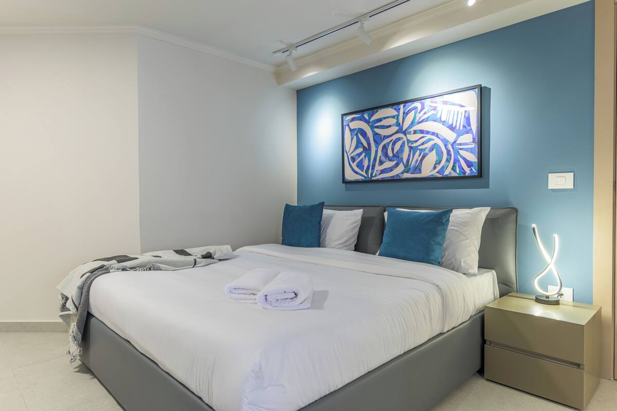 Un dormitorio moderno con una cama grande, almohadas azules y blancas, una obra de arte en la pared y una mesita de noche con lámpara.
