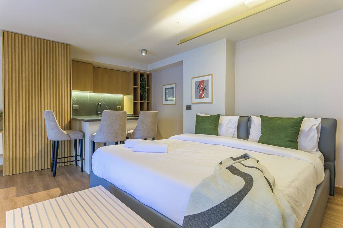 Habitación de hotel moderna con una cama grande, dos taburetes altos, una kitchenette pequeña y un separador de madera.