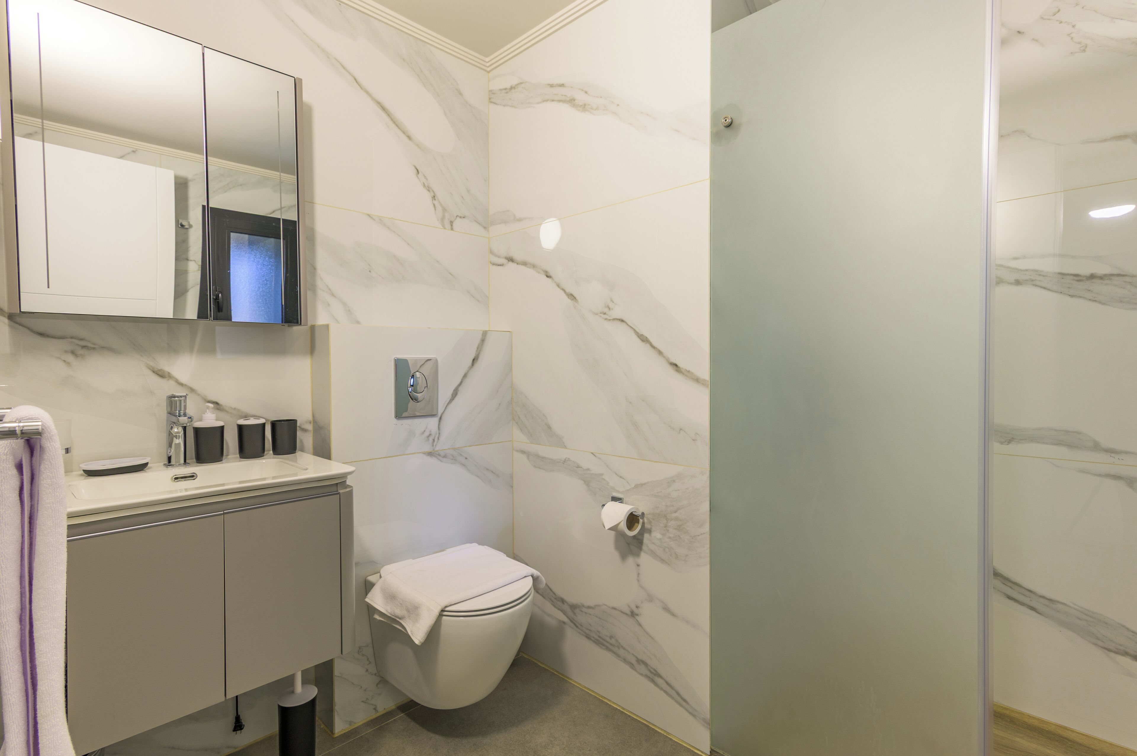 A bathroom with a marble wall, a toilet, a sink with a mirror, and a shower area.