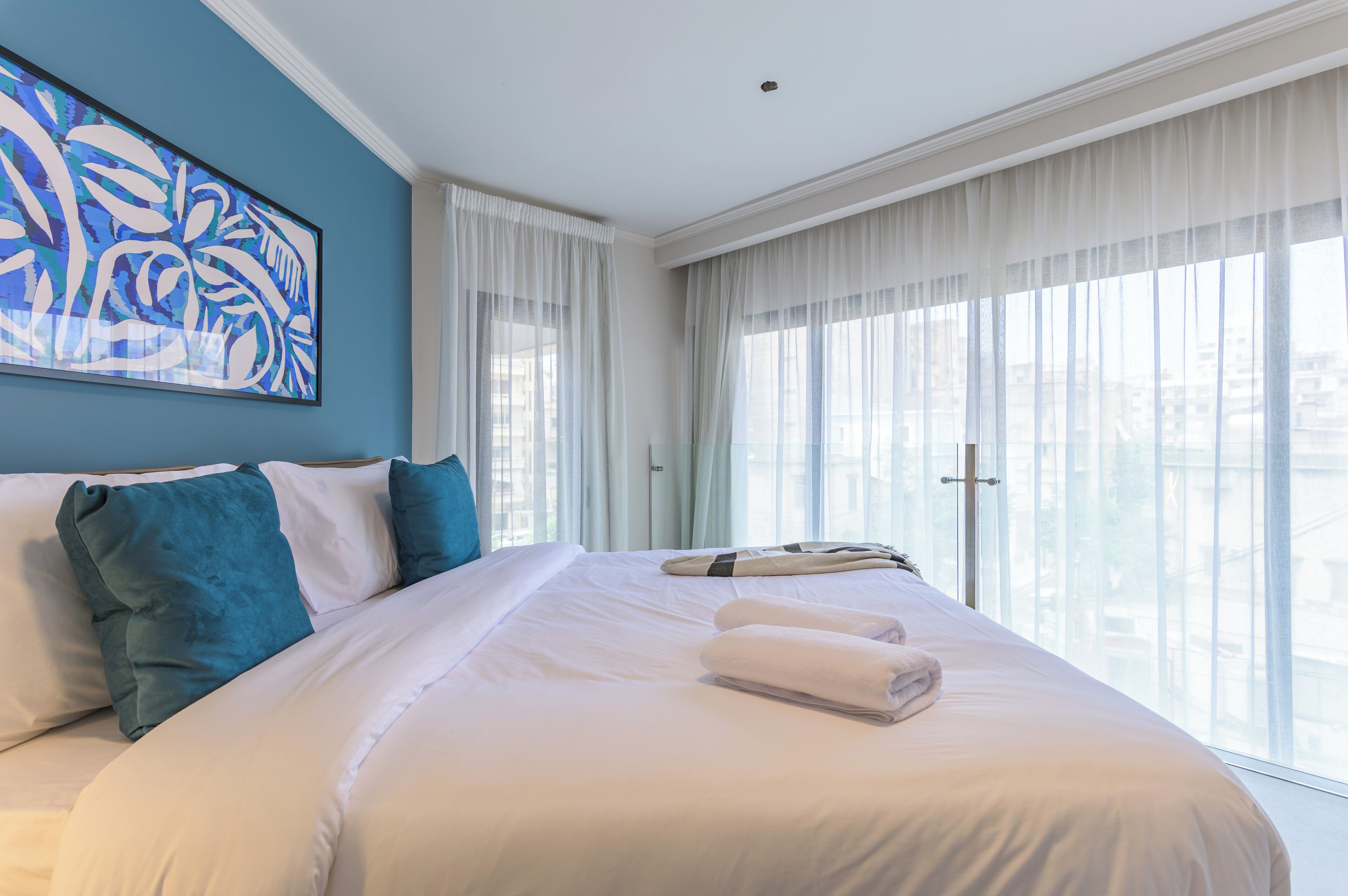 A hotel room with a large bed, white bedding, teal pillows, and a city view through sheer curtains.