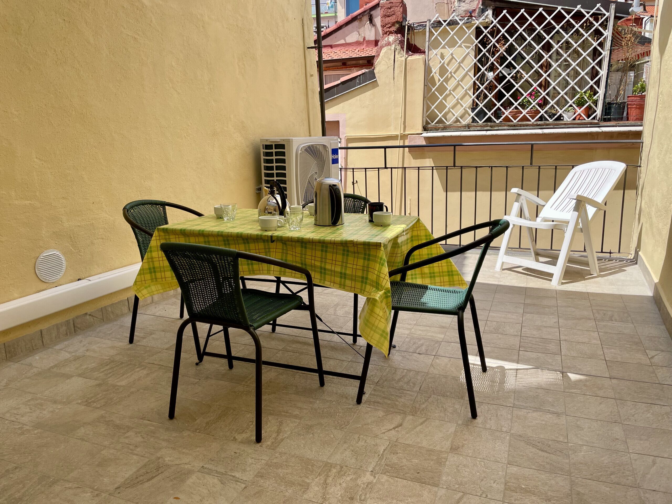 A small outdoor patio with a table covered by a yellow checkered tablecloth, two green metal chairs, and a white plastic chair.
