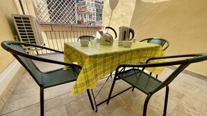 Un balcón con una mesa cubierta por un mantel a cuadros amarillo, cuatro sillas de mimbre verde y una baranda de metal.