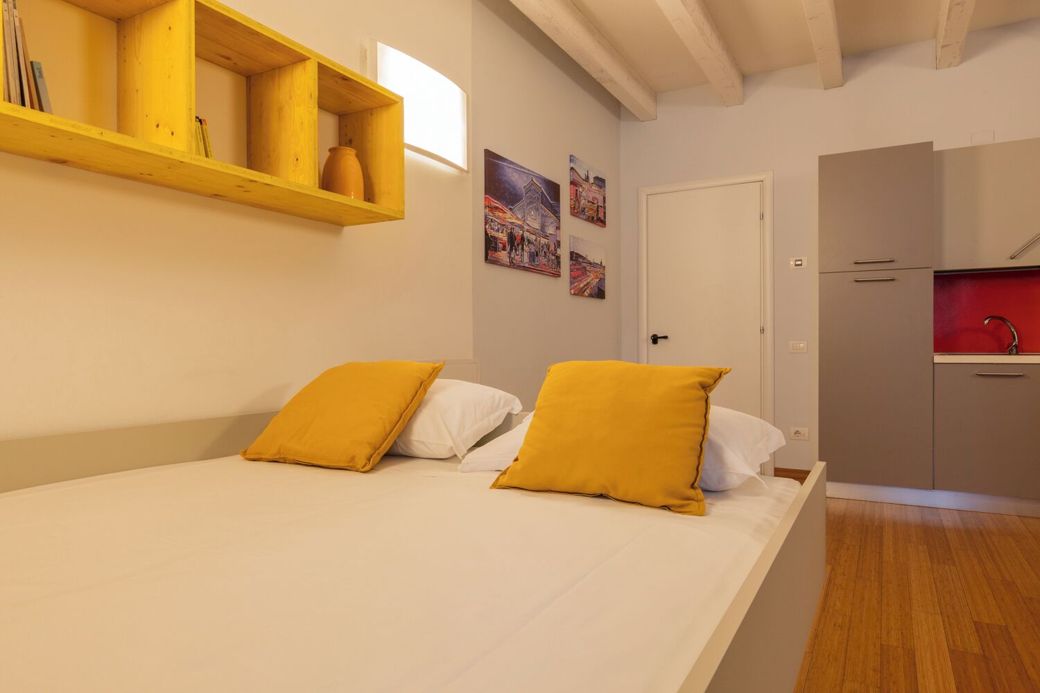 A bedroom with a bed, two pillows, a wooden shelf with a vase, and a door leading to a kitchen.