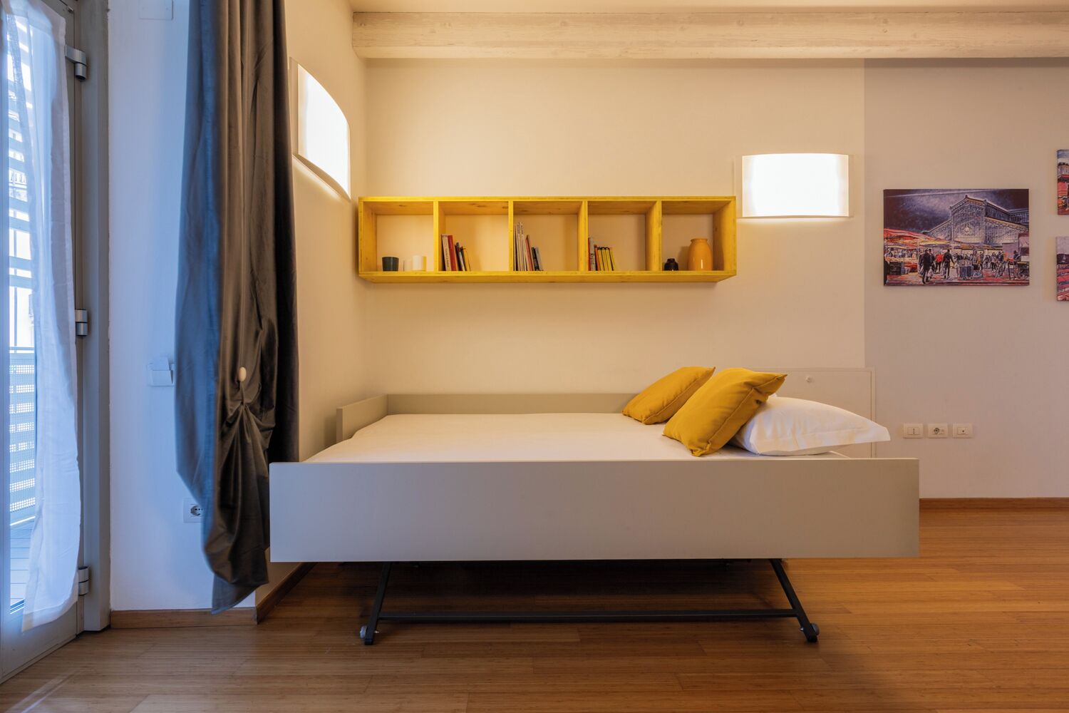 Una habitación moderna y minimalista con un sofá cama blanco, una estantería de madera con libros y un cuadro en la pared.