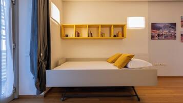 Una habitación moderna y minimalista con un sofá cama blanco, una estantería de madera con libros y un cuadro en la pared.