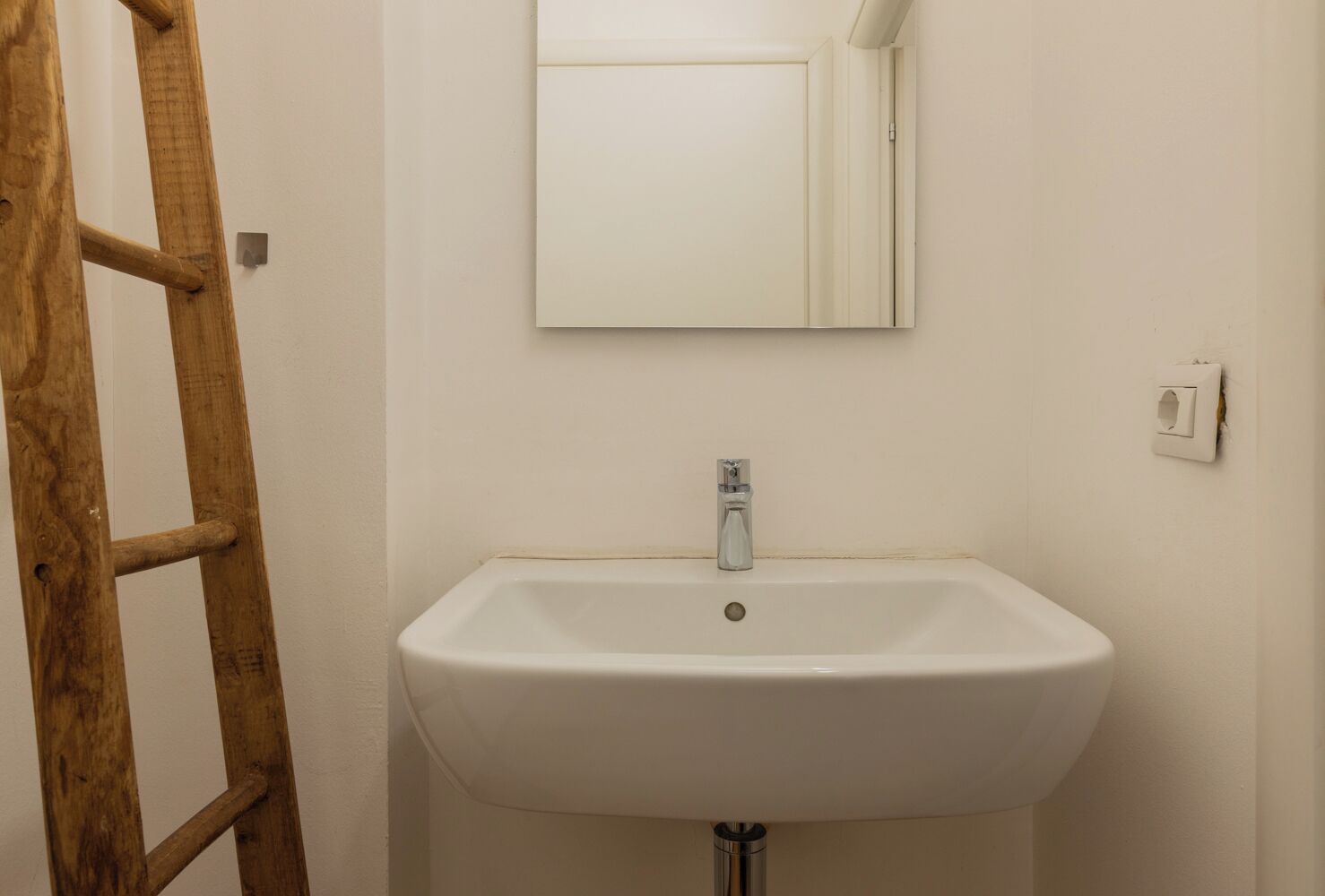 A bathroom with a white sink, a mirror above it, and a wooden ladder leaning against the wall.