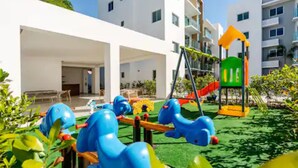 Zona de juegos infantiles con tobogán y estructuras para escalar, rodeada por un edificio con balcones y un área de estar techada.