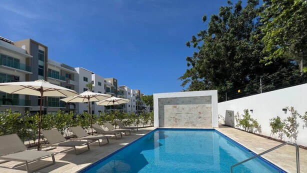 Un área de piscina moderna con sillas de descanso y sombrillas, rodeada de edificios y follaje.