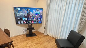 A living room with a flat-screen TV displaying a streaming service interface, a wooden dining table, a black sofa, and a window with sheer curtains. - One private house! Max. 9 people with parking! 3 min walk from Yoyogi, 2 min by train from Shinjuku/Harajuku, 5 min from Shibuya, 7 min from Roppongi Hotel Like Yoyogi Homes (Sibuya-ku)