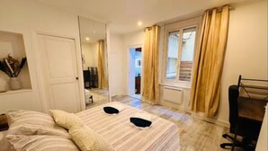Un dormitorio con cama, escritorio y silla. Hay una ventana con cortinas, una puerta y una pequeña mesa con un florero.