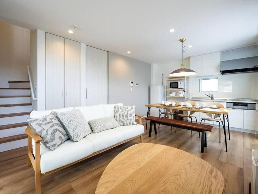 A modern apartment with a white sofa, wooden dining table, and a kitchen area with white cabinets and a microwave.