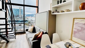 Ein modernes Wohnzimmer mit großem Fenster, einer Couch, einem Esstisch und einer Treppe.
