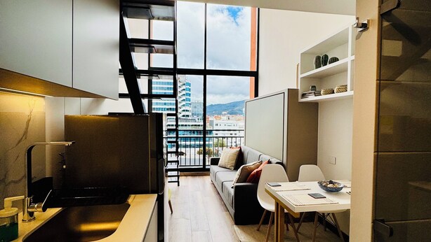 Eine moderne Küche mit integriertem Spülbecken, ein Essbereich mit Tisch und Stühlen, eine Couch und Blick auf Gebäude durch große Fenster.