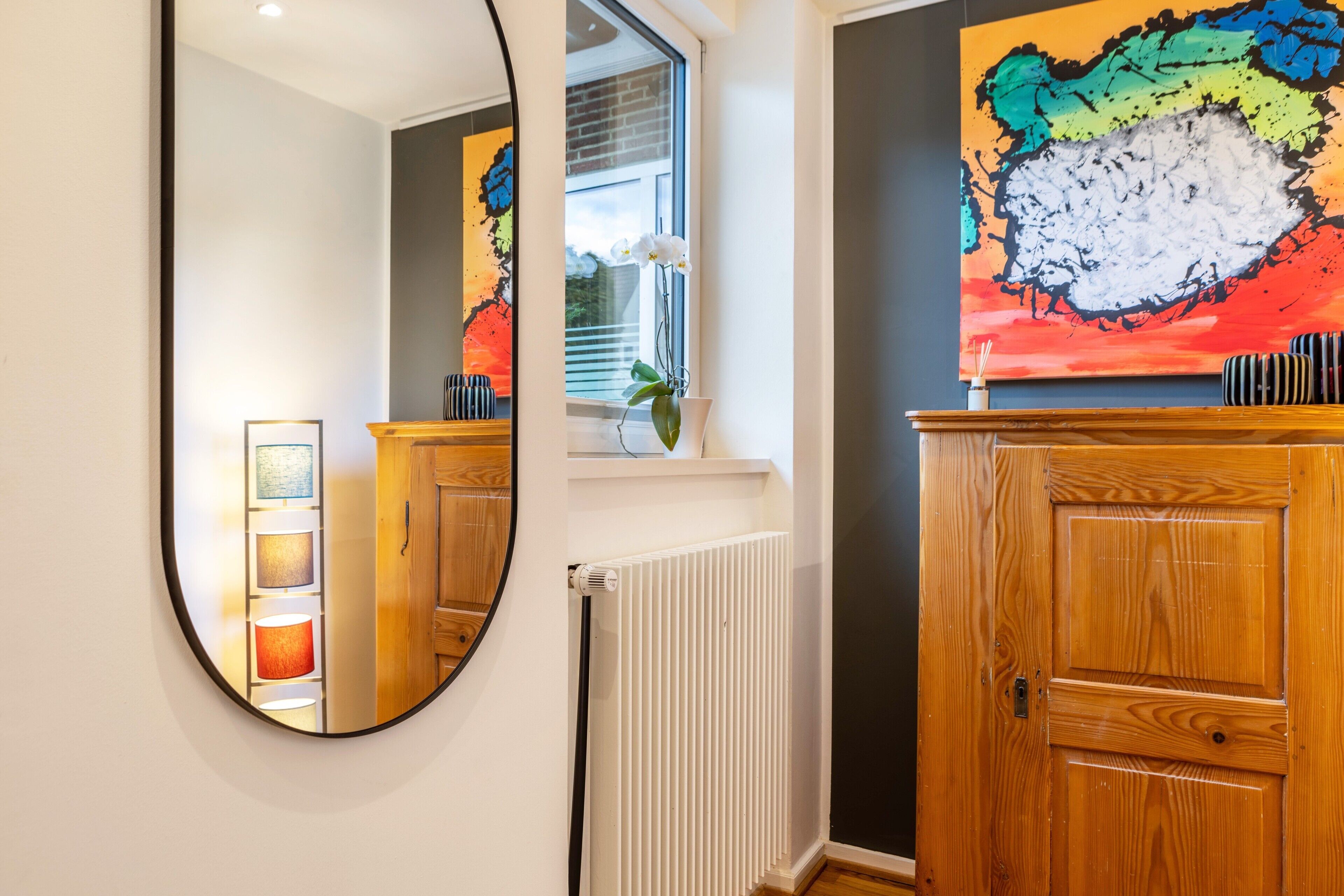 Een badkamer met een houten kast, een spiegel, een radiator en een kleurrijk abstract schilderij aan de muur.