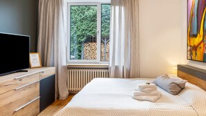 Un dormitorio con cama, televisor, ventana con cortinas, radiador y un cuadro en la pared.