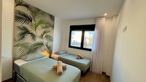 A room with two beds, a large window with a view, and a tropical leaf mural on the wall.