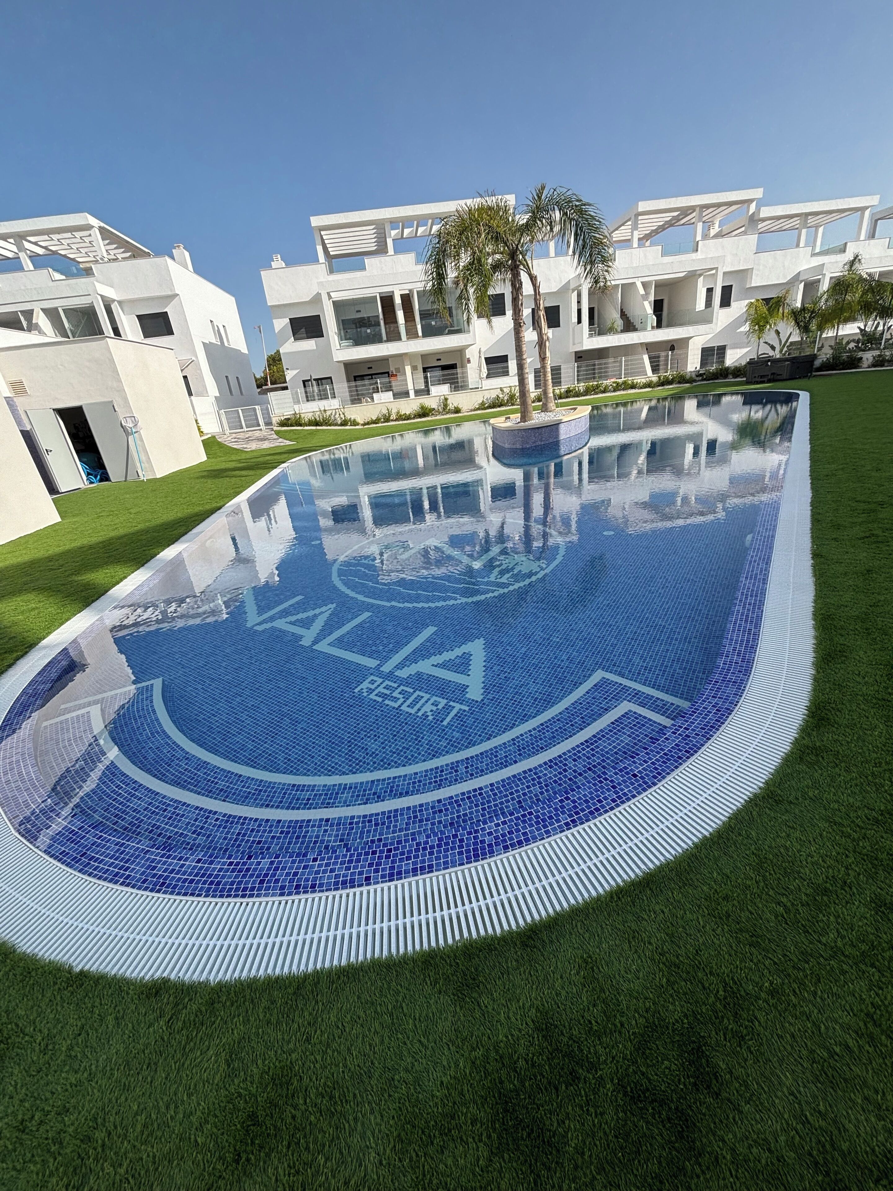 Een modern zwembad met een helderblauw mozaïekpatroon, omgeven door verzorgd gras en palmbomen, met moderne gebouwen op de achtergrond.