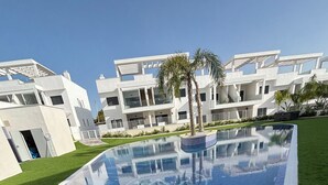 A modern pool with a clear blue mosaic design, surrounded by well-maintained grass and palm trees, with contemporary buildings in the background.