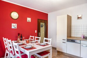 Un comedor con pared roja, mesa y sillas blancas, un reloj, y una cocina con armarios blancos y encimera de madera.