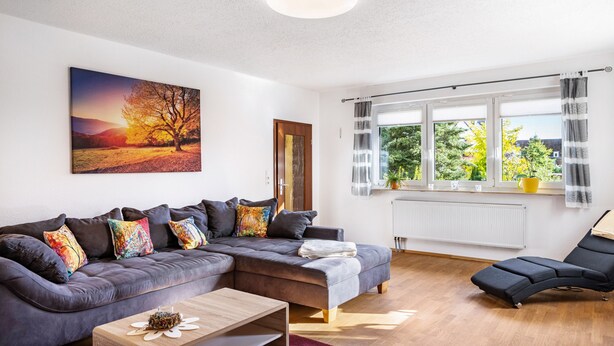 Un salon moderne avec un canapé d’angle, un fauteuil, une table basse et une grande fenêtre avec des rideaux.