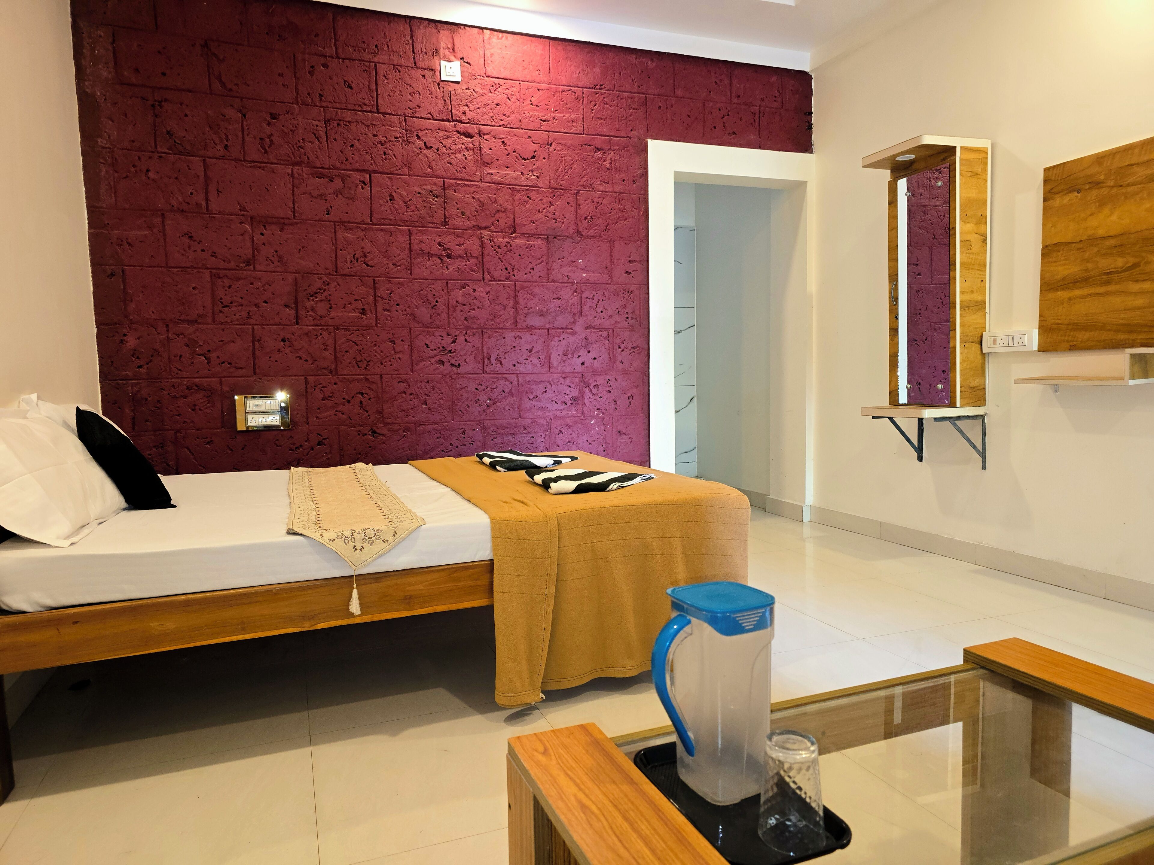 Um quarto com parede de tijolos, uma cama com manta bege, uma mesa de cabeceira com jarra de água e um espelho na parede.