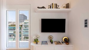 A modern living room with a large TV mounted on the wall, a white console table with decorative items, and a view of a high-rise building through the glass doors. - Michalakopoulou Corner & Bright Apartment, Luxury (Athens)