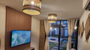 Un salon moderne avec un téléviseur fixé sur un panneau en bois, deux lustres suspendus, un canapé et une vue sur un mur de briques et une porte en verre donnant sur l’extérieur.