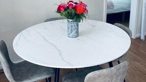 A round dining table with a marble top, surrounded by four grey upholstered chairs with black legs. A vase with red and pink flowers is placed on the table. A round mirror with a wooden frame hangs on the wall above the table. - Ivy Cottage Two Bedroom & Garden in Temple Fortune (London)