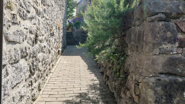 A narrow cobblestone alley flanked by stone walls and greenery, leading to a building with a visible staircase.