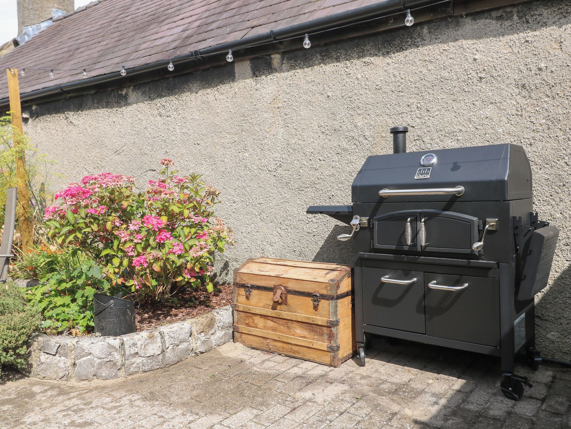 A barbecue grill with a storage chest and a potted plant with pink flowers against a textured wall.