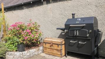 A barbecue grill with a storage chest and a potted plant with pink flowers against a textured wall.